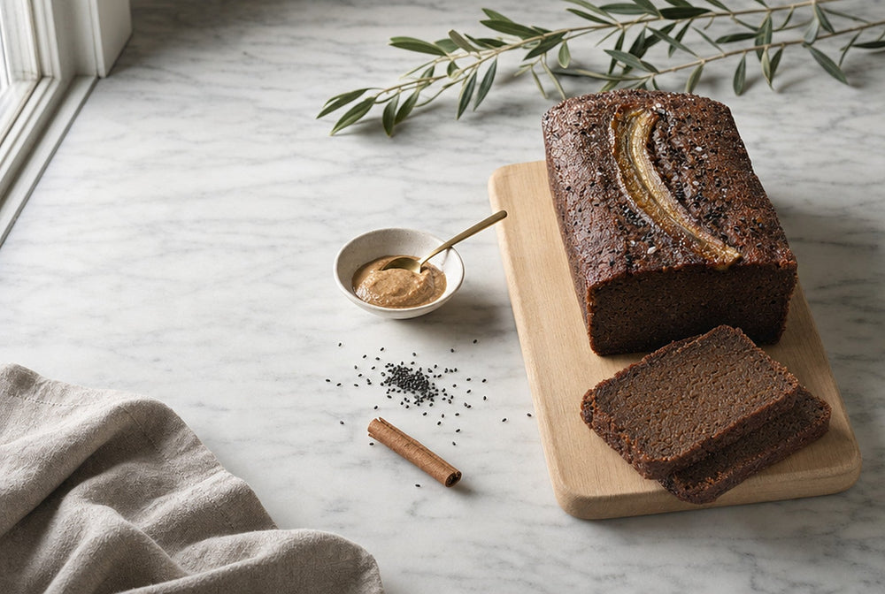 A flourless roasted-banana loaf on a pale wooden board on marble, two thick slices cut, with almond butter, black sesame, a cinnamon quill and an olive branch