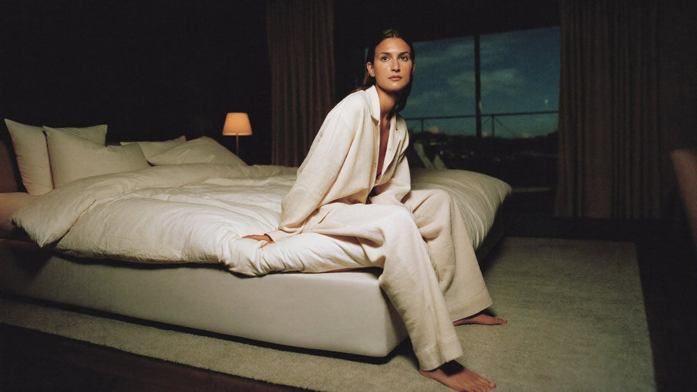 A woman in cream linen pyjamas sits on the edge of her bed before dawn in September, the window still dark behind her